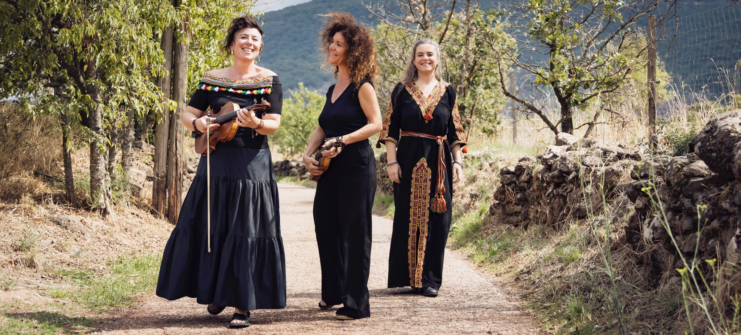 Trois musiciennes marchent en riant sur un chemin de campagne, violons à la main